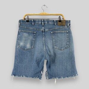 Może przedstawiać: Kr&oacute;tkie spodenki dżinsowe w kolorze niebieskim z frędzlami na dole i brązową sk&oacute;rzaną metką Wrangler na tylnej kieszeni.