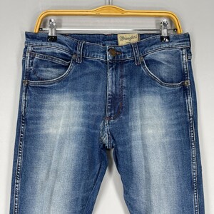 May include: Blue denim jeans with a faded wash, featuring the "Wrangler" logo on the button. The jeans are hung on a wooden hanger. The jeans have classic five-pocket styling.