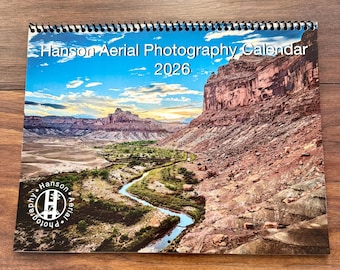 May include: A spiral-bound calendar titled "Hanson Aerial Photography Calendar 2026" featuring a scenic landscape photograph. The image showcases a winding river through a canyon with red rock formations under a blue sky.