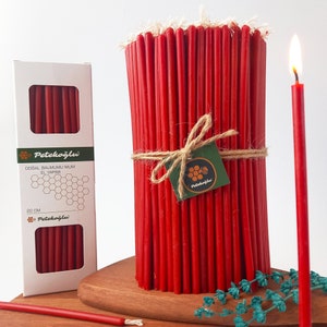 May include: A bundle of 20 cm red beeswax candles, tied with twine and a small label that reads "Petekoglu". The candles are arranged in a cylindrical shape and are displayed on a wooden surface. The image also shows a single red beeswax candle.