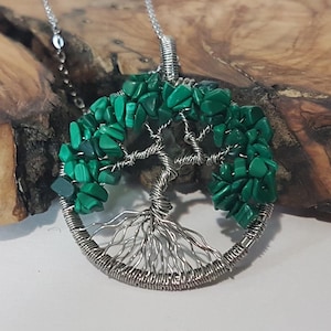 Puede incluir: Un colgante de árbol de la vida envuelto en alambre plateado con piedras de malaquita verde. El colgante está en una cadena plateada y cuelga de una superficie de madera.
