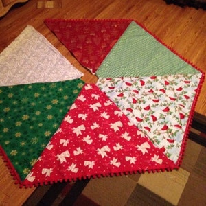 May include: A Christmas tree skirt made of fabric triangles in various patterns, including red with white angels, green with white snowflakes, and white with a silver pattern. The skirt has a red pom-pom trim.