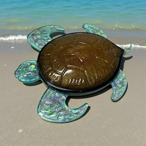 May include: A decorative turtle figurine with a brown, textured shell and iridescent blue-green flipper-like appendages. The turtle is set on a sandy beach with the ocean in the background. The shell has a black border.