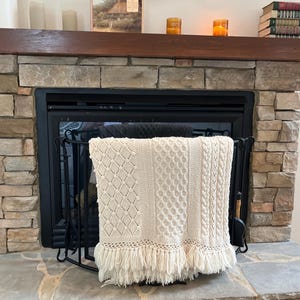 May include: An off-white knitted throw blanket featuring a variety of textured patterns including diamond, honeycomb, and cable knit designs. The rectangular blanket is finished with a long, thick fringe along the bottom edge and is draped over a black metal fireplace screen.
