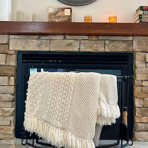 May include: A cream-colored knit throw blanket featuring a variety of textured patterns including cables, diamonds, and bobbles. The blanket is finished with long, thick fringe along the bottom edge and is draped over a black metal fireplace screen.