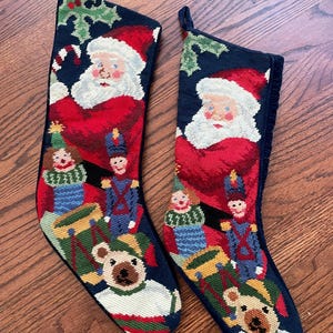 Puede incluir: Dos calcetines navideños con fondo azul marino. Cada calcetín presenta un diseño de Papá Noel, junto con soldados de juguete, un oso de peluche y otros elementos festivos. Los calcetines están hechos de material de punto de aguja.