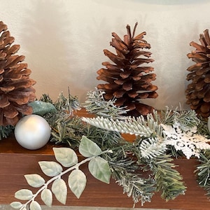 Pode incluir: Uma guirlanda festiva com três pinhas grandes castanhas, dois ornamentos esféricos prateados mate, agulhas de pinheiro artificiais verdes com acabamento branco geado, folhas verdes e um ornamento de floco de neve branco recortado.