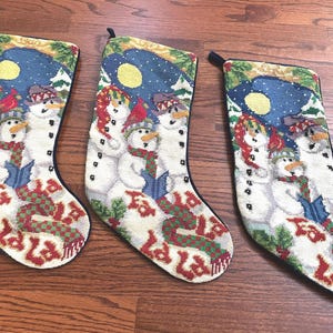 Puede incluir: Tres calcetines navideños con diseño de muñeco de nieve. Cada calcetín presenta muñecos de nieve con sombreros y bufandas, cantando villancicos bajo un cielo estrellado. La palabra "La La La" está bordada en rojo y verde.