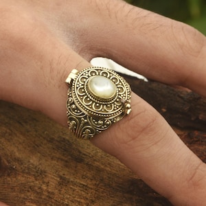 May include: A gold ring with an ornate design and a white pearl-like stone in the center. The ring is openable and has a hinged lid.