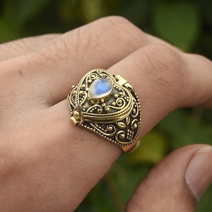 May include: A gold heart-shaped ring with a hinged lid and a moonstone set in the center. The ring is detailed with intricate floral designs.