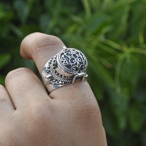 May include: A silver ring with a round, filigree-detailed compartment that opens. The ring is worn on a finger.
