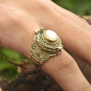 May include: A gold ring with an ornate design and a white pearl in the center. The ring is a hinged locket that opens to reveal a small compartment.