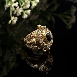 May include: A gold-colored ring with intricate floral and scrollwork designs. The ring features a large, oval-shaped black gemstone at the center. The ring is highly detailed and appears to be made of metal.