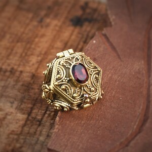 May include: Ornate gold-colored hexagonal pendant with a hinged lid, featuring intricate scrollwork and a central oval, deep red gemstone. The pendant is set on a textured, brown surface.