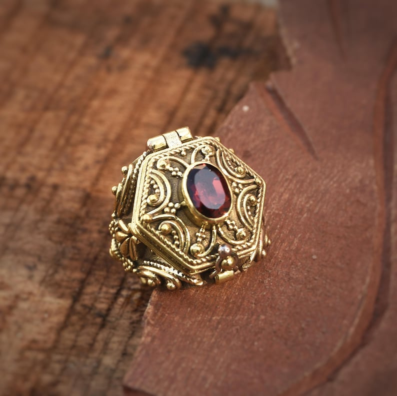 May include: Ornate gold-colored hexagonal pendant with a hinged lid, featuring intricate scrollwork and a central oval, deep red gemstone. The pendant is set on a textured, brown surface.