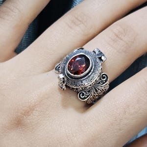 Op de afbeelding: Oogverblindende zilveren ring met een grote, ovale, dieprode edelsteen. De ring heeft ingewikkelde filigraan- en krulwerkdetails rond de steen en op de band. Een opvallend sieraad voor speciale gelegenheden.