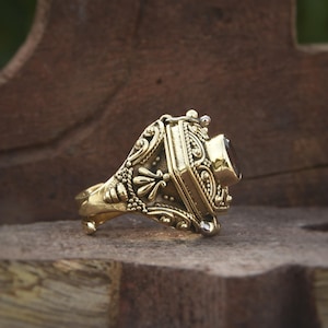 May include: Ornate gold-colored ring with a rectangular, dark gemstone. The ring features intricate floral and scrollwork designs along the band and setting. The gemstone is set in a rectangular frame, adding to the ring's vintage aesthetic.