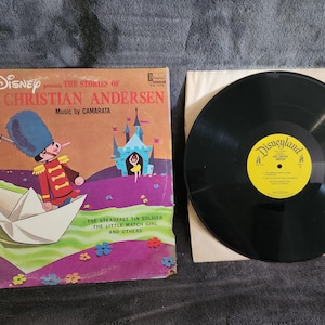 May include: A vintage Walt Disney record album featuring the stories of Hans Christian Andersen. The album cover features a colorful illustration of a toy soldier on a paper boat, a castle, and a little girl. The album title is printed in bold, red letters. The record is in a white paper sleeve with the Disneyland logo printed on it.