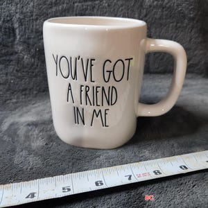 Op de afbeelding: Een witte keramische mok met de tekst "YOU'VE GOT A FRIEND IN ME" in zwarte letters. De mok heeft een gebogen handvat en staat tegen een grijze achtergrond. Een liniaal is bij de mok geplaatst.