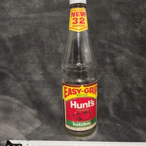Puede incluir: Una botella de vidrio de ketchup Hunt's con una etiqueta roja y una etiqueta amarilla Easy-Grip bottle. La botella es de 32 onzas y tiene una tapa blanca.