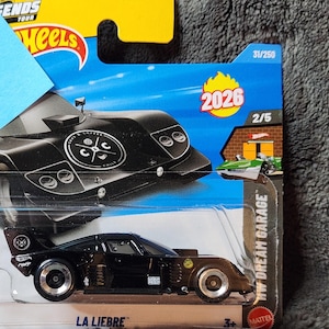 May include: A black Hot Wheels die-cast car, "La Libre," with silver wheels and a distinctive fin. The packaging includes the "HW Dream Garage" logo, the number 31/250, and the year 2026. The car features the logo of "Cars & Coffee" on the top.