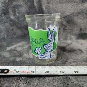 Op de afbeelding: Een helder drinkglas met een blauwe lijntekening van Bugs Bunny die een wortel vasthoudt tegen een groene achtergrond. Het glas staat naast een wit meetlint, waaruit blijkt dat het glas een diameter heeft van ongeveer 7,6 cm.