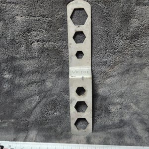 Puede incluir: Una llave de metal plateado con múltiples aberturas hexagonales de varios tamaños. La palabra "VICTOR" está estampada en la herramienta. La llave se muestra sobre una superficie gris texturizada con una cinta métrica en primer plano.