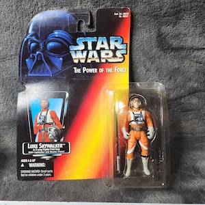 Op de afbeelding: Een Star Wars "The Power of the Force" actiefiguur van Luke Skywalker in X-wing Fighter Pilot Gear. De figuur zit in een doorzichtige plastic hoes die aan een kaart is bevestigd met het Star Wars-logo en de helm van Darth Vader. De figuur is ongeveer 7.6 cm groot.