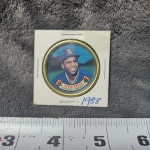 Może przedstawiać: Karta baseballowa w stylu vintage z Alvinem Davisem z Seattle Mariners. Karta ma okrągły obraz Davisa w jego mundurze Mariners, umieszczony w złotej ramce. Rok "1988" jest wydrukowany poniżej.