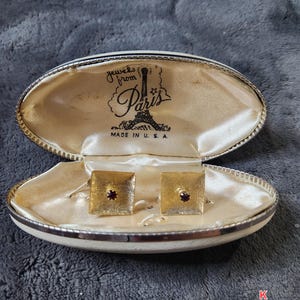 May include: Open oval jewelry case with gold-colored square cufflinks. Each cufflink has a small, red gemstone in the center. The case is lined with cream-colored satin and has the text "jewels from Paris" and "MADE IN U. S. A."