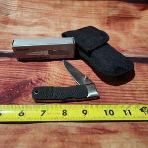 May include: A black lockback knife with a silver blade and a black handle. The knife is closed and is sitting on a wooden surface. The knife is in a black nylon sheath. The knife is labeled "Maxam Lockback Knife" on the box.