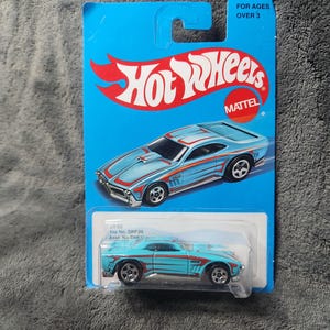 May include: A Hot Wheels toy car on a blue and white card. The car is light blue with red stripes and silver wheels. The card reads "Hot Wheels" and "MATTEL". The packaging is marked "FOR AGES OVER 3".