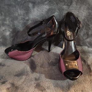 Può includere: Paio di sandali neri con tacco alto e cinturini alla caviglia. Le scarpe a punta aperta presentano un tessuto a fantasia rosa e nero sulla tomaia e un tacco nero lucido. Il marchio "Rock & Republic" è visibile sulla soletta.