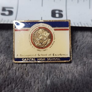 May include: Rectangular pin with a gold border. The pin features a cream-colored background with text and a gold emblem. The top has a blue stripe with white stars. The bottom has a blue stripe with the words "CAPITAL HIGH SCHOOL".