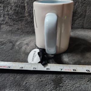 K&ouml;nnte beinhalten: Eine wei&szlig;e Keramiktasse mit hellblauem Henkel und Innenraum. Die Tasse hat schwarzen Text und ein Etikett mit der Aufschrift "Frozen". Die Tasse ist etwa 10 cm hoch.