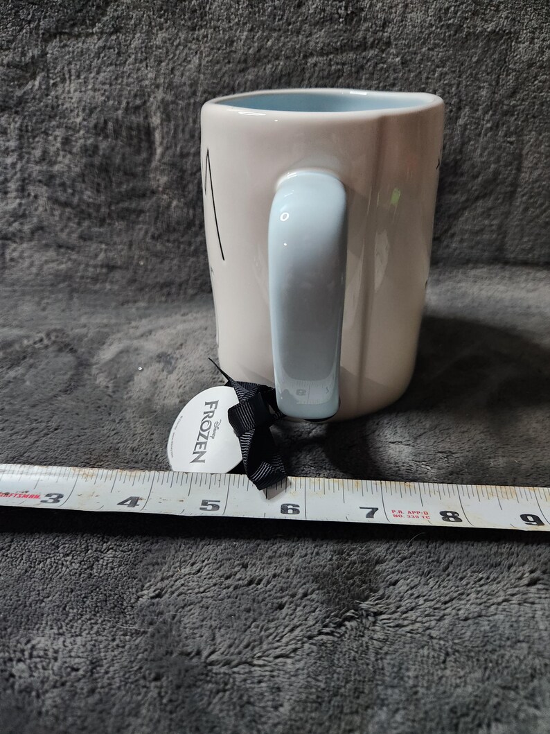 K&ouml;nnte beinhalten: Eine wei&szlig;e Keramiktasse mit hellblauem Henkel und Innenraum. Die Tasse hat schwarzen Text und ein Etikett mit der Aufschrift "Frozen". Die Tasse ist etwa 10 cm hoch.