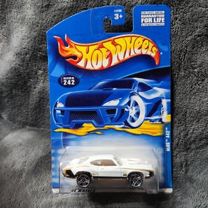 May include: A white and gold Hot Wheels die-cast car, OLDS 442 model, in its original blue packaging. The packaging features the Hot Wheels logo, a flame graphic, and the text "DIE-CAST METAL GUARANTEED FOR LIFE & PLASTIC PARTS".