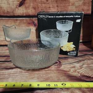 以下が含まれることがあります： テクスチャ加工された表面の、3ピースの透明ガラス製サービングセット。セットには、2つのボウルと、金属製のスタンドが付いた小さなボウルが含まれています。箱には「CRISTALLIN Service à croustilles et trempette 3 pièces」と書かれています。