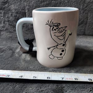K&ouml;nnte beinhalten: Wei&szlig;er Keramikbecher mit hellblauem Innenraum und Griff. Der Becher zeigt eine schwarze Strichzeichnung von Olaf aus Frozen. Ein schwarzes Etikett ist am Griff befestigt. Der Becher ist etwa 10 cm hoch.