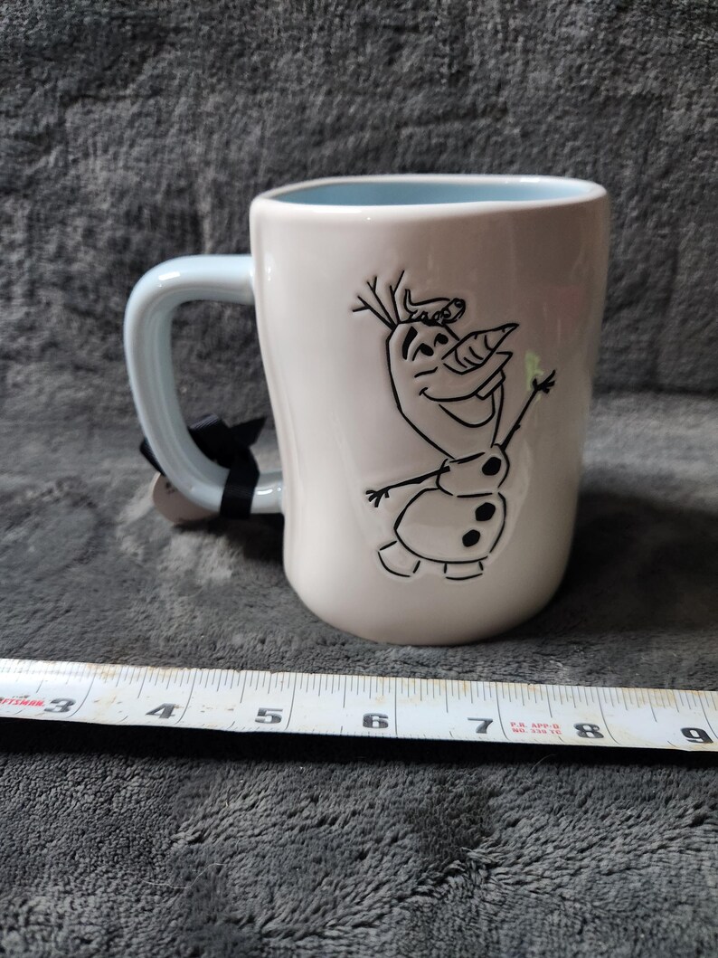 K&ouml;nnte beinhalten: Wei&szlig;er Keramikbecher mit hellblauem Innenraum und Griff. Der Becher zeigt eine schwarze Strichzeichnung von Olaf aus Frozen. Ein schwarzes Etikett ist am Griff befestigt. Der Becher ist etwa 10 cm hoch.