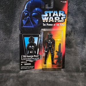 May include: A Star Wars action figure of a TIE Fighter Pilot in black and grey with a black helmet and a blaster pistol and rifle. The figure is packaged in a plastic blister pack with a red, yellow, and blue background. The packaging includes the text "TIE FIGHTER PILOT" and "Imperial Blaster Pistol and Rifle!"  The packaging also includes a warning about choking hazards for children under 3 years old.