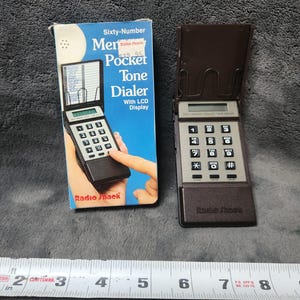 May include: A vintage Radio Shack Sixty-Number Pocket Tone Dialer with LCD display. The brown and gray device is open, next to its original box. A measuring tape is in the foreground.