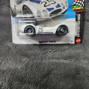 May include: A white Hot Wheels Maserati Tipo 61 die-cast toy car with the number 25 printed on the side. The car is enclosed in its original blister pack packaging, which features a graphic of the same car and the text 2026 and 9/10.