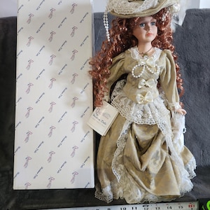 May include: A porcelain doll wearing a white lace dress with a gold pattern, a white lace hat with a white flower, and a pearl necklace. The doll has long red curly hair and blue eyes. The doll is standing on a white surface with a measuring tape in the foreground.