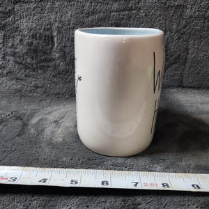 K&ouml;nnte beinhalten: Wei&szlig;er Keramikbecher mit hellblauem Innenraum. Der Becher hat einen schwarzen Schriftzug &bdquo;hello&ldquo; und ein Blumendesign. Der Becher ist etwa 10 cm hoch.