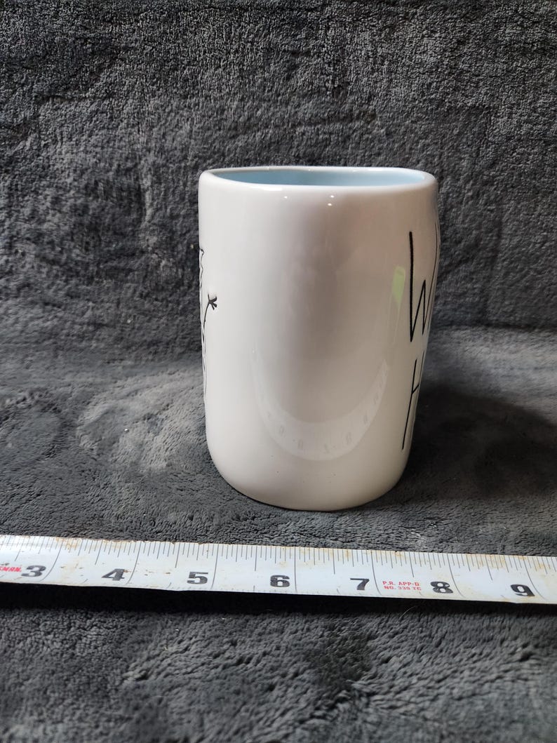 K&ouml;nnte beinhalten: Wei&szlig;er Keramikbecher mit hellblauem Innenraum. Der Becher hat einen schwarzen Schriftzug &bdquo;hello&ldquo; und ein Blumendesign. Der Becher ist etwa 10 cm hoch.