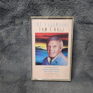 May include: A vintage cassette tape of "The Essential Tom T. Hall: Twentieth Anniversary Collection." The cover features a portrait of Tom T. Hall against a backdrop of a sunset over a landscape. The cassette is in a clear plastic case.