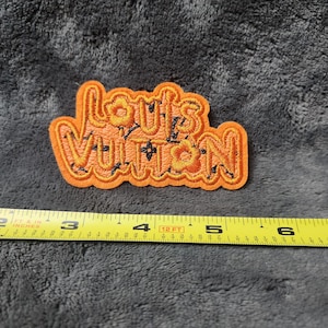 Peut inclure: Un écusson brodé orange portant le texte Louis Vuitton dans une police stylisée avec des accents floraux noirs. Un mètre ruban jaune est placé sous l'écusson, indiquant une largeur d'environ 11,5 cm pour le motif brodé.
