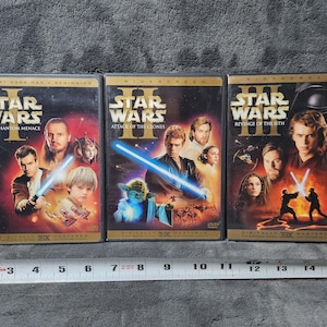 May include: Three Star Wars DVD movie cases. The Phantom Menace, Attack of the Clones, and Revenge of the Sith are displayed. Each case features character images and the Star Wars logo. The cases are in a row, with a measuring tape below.