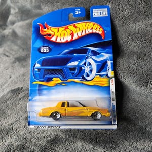 May include: A yellow Hot Wheels die-cast car in its original packaging. The packaging is blue with the Hot Wheels logo and the text "GUARANTEED FOR LIFE". The car is a Montezuma model from the 2001 First Editions series.